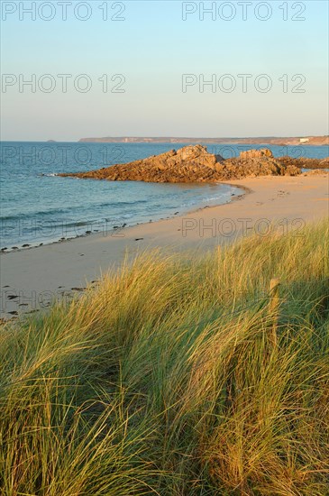 A tranquil beach scene with grassy dunes in the foreground leading to a rocky outcrop by the sea, Erquy