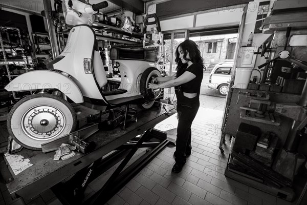 Woman mechanic working on repairing a white moped italian vintage scooter, surrounded by tools in a workshop, real women performing traditional man jobs of the past, black and white photograph