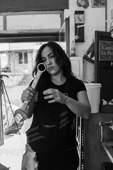 Woman mechanic holding a large wrench, examining it thoughtfully in a workshop, real women performing traditional man jobs of the past, black and white photograph