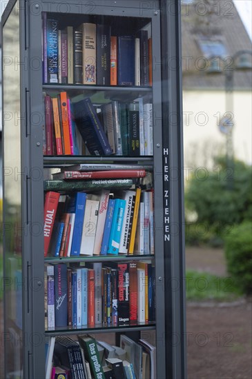 Open bookcase, close-up, many colourful books in a glass case, outdoor, in the background blurred lantern, green bushes and a house, Volmarstein, Ruhr area, Germany, Europe