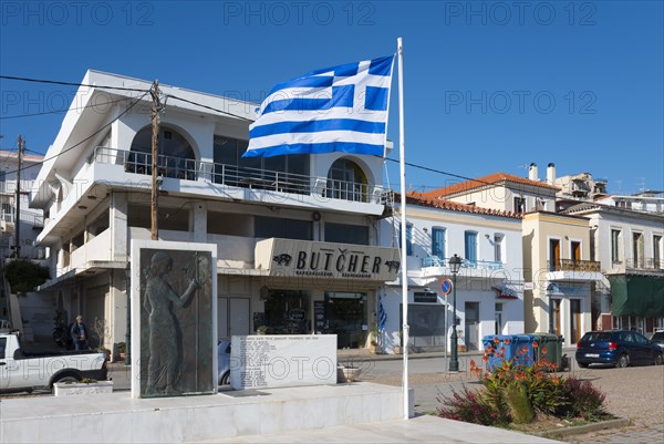 Greek flag in front of a building with the inscription 'BUTCHER' and a statue sculpture, Galatas, Argolis, Peloponnese, Greece, Europe