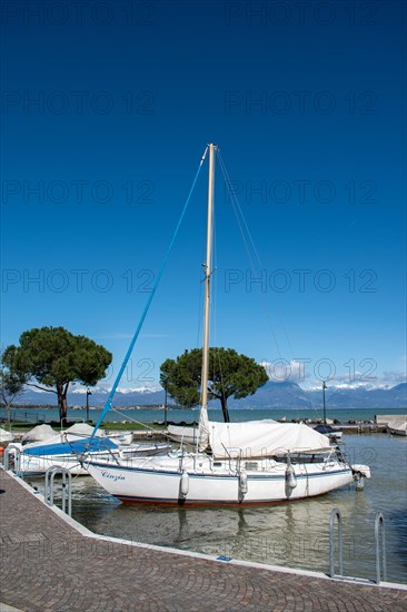 A sailing boat moored in the harbour with clear blue sky and mountain in the background, Sirmione, Lake Garda, Italy, Europe