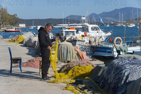 A man repairs traditional fishing nets at the sunny marina with boats and mountains in the background