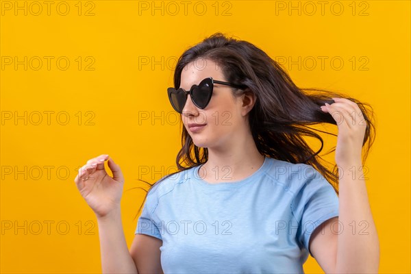 Studio portrait with yellow background of a caucasian adult woman wearing heart shape sunglasses arranging her hair