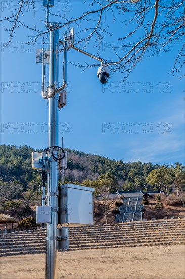 Wi-fi, CCTV with 5 G transmitters on chrome pole in from of evergreen tree in public park with lake and blue sky in background in South Korea