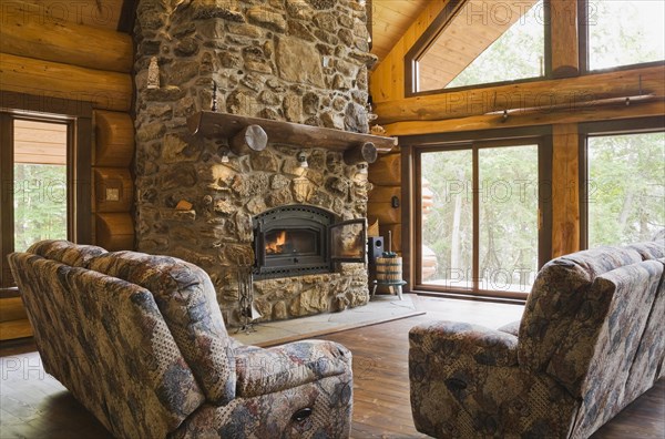 Upholstered sofas with lit fieldstone and porous rock fireplace in living room inside handcrafted red cedar log cabin home, Quebec, Canada, North America