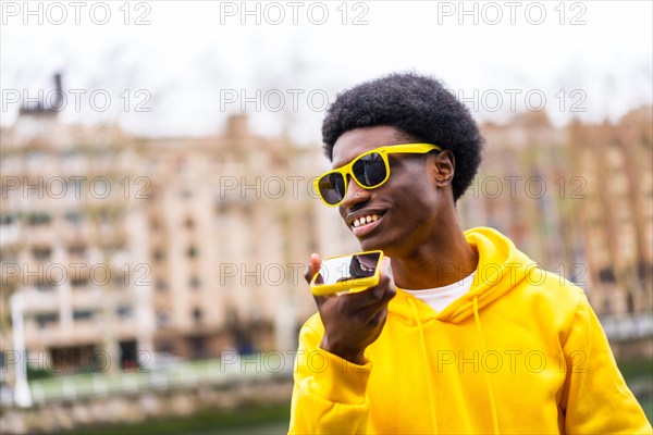 Cool african young man sending voice message with mobile standing in the city