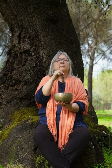 Mature woman with white hair in her sixties meditating in the forest with her eyes closed and a Tibetan singing bowl ringing