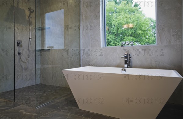 Clear glass shower stall and white freestanding vessel shaped bathtub in bathroom with grey ceramic tile floor and walls on ground floor inside modern cubist style home, Quebec, Canada, North America