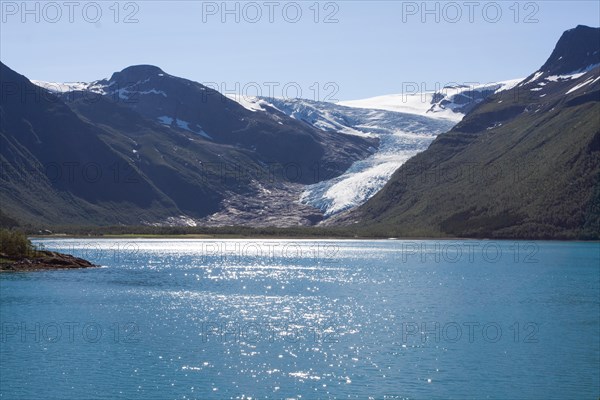 A glacier flows between snow-capped mountains into a clear blue lake under bright sunshine Arctic Ocean Lofoten