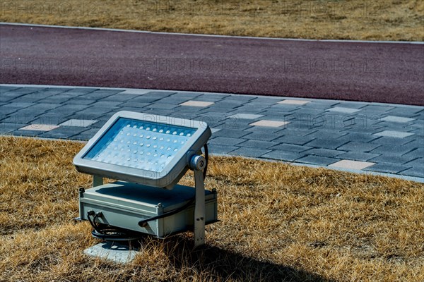 An LED floodlight installed on grass next to a paved path in daylight, in South Korea