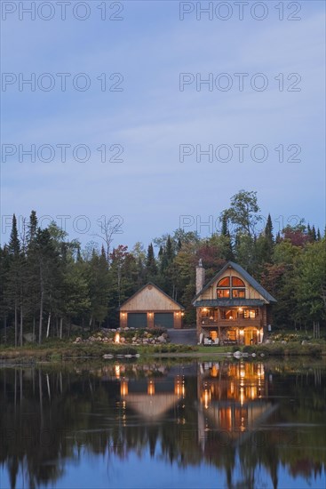 Illuminated two car garage and handcrafted two story spruce log cabin home with fieldstone chimney and green sheet metal roof on edge of lake at dusk in autumn, Quebec, Canada, North America