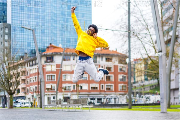 Cool african man jumping while listening to music using headphones in the city