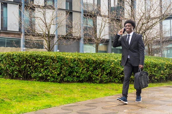 African young businessman commuting while talking to the mobile outdoors along a public urban park