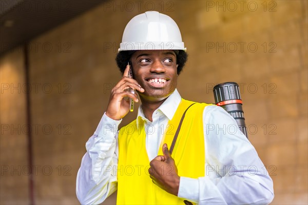 Portrait of a happy young african architect wearing protective work clothes and helmet talking to the phone while visiting construction site