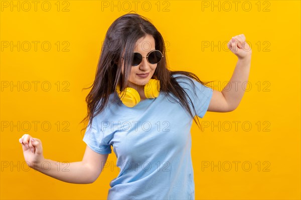 Studio portrait with yellow background of happy woman dancing wearing sunglasses and headphones on the neck