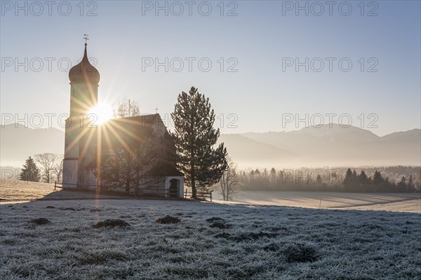 Chapel at sunrise in front of mountains, backlight, sunbeams, hoarfrost, winter, Sankt Johannisrain, Alpine foothills, Bavaria, Germany, Europe