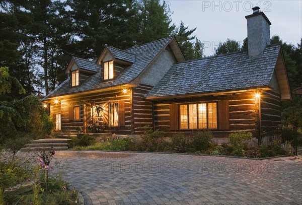 Illuminated old 1800s reconstructed log cabin home facade and paving stone driveway at dusk, Quebec, Canada, North America
