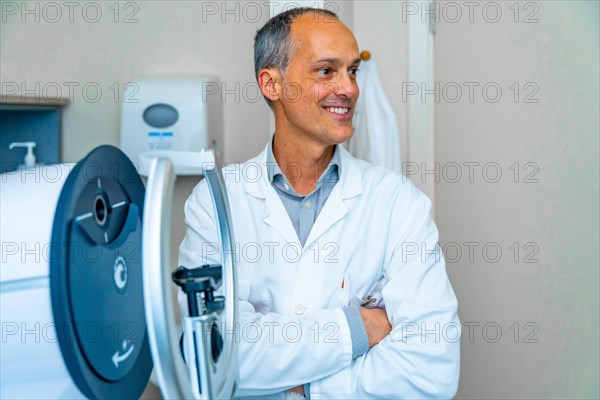 Proud mature male ophthalmologist looking distracted next to an innovative laser machine in a clinic