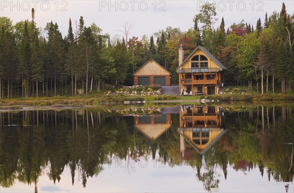 Garage and handcrafted two story spruce log cabin home with fieldstone chimney and green sheet metal roof on edge of lake in autumn, Quebec, Canada, North America