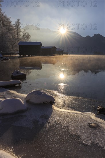 Evening mood at mountain lake in front of mountains, boat huts, shore, winter, snow, reflection, sunbeams, backlight, Lake Kochel, Alpine foothills, Bavaria, Germany, Europe
