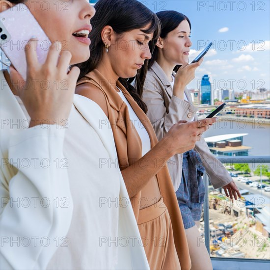 Three women are standing on a balcony, each holding a cell phone. They are all dressed in business attire, and the scene suggests a moment during a busy day