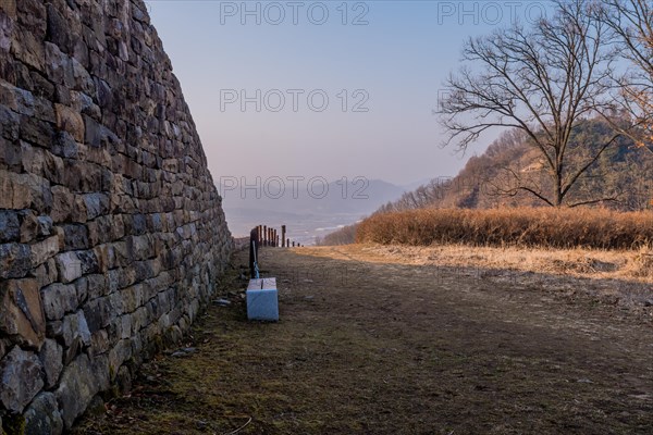 Park bench beside mountain fortress wall made of flat stones with hazy blue sky in background in Boeun, South Korea, Asia