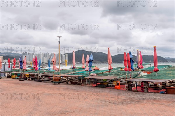 Folded beach umbrellas line an empty waterfront against a cloudy sky with distant cityscape, in Ulsan, South Korea, Asia