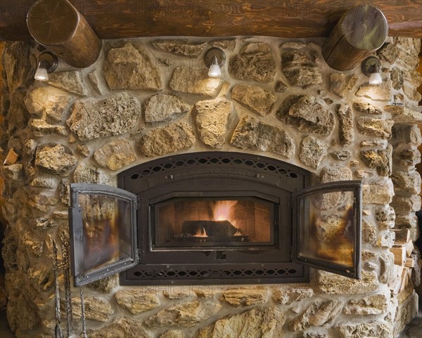 Lit fieldstone and porous rock fireplace in living room inside handcrafted red cedar log cabin home, Quebec, Canada, North America