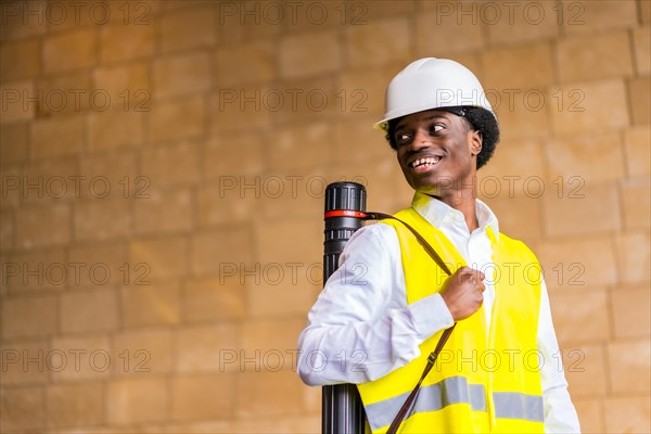 Horizontal photo with copy space and low angle view of a proud african architect wearing helmet and waistcoat holding a drawing tube in the city