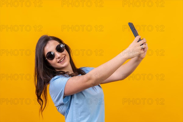 Studio portrait with yellow background of a chic woman with sunglasses taking a selfie smiling at camera