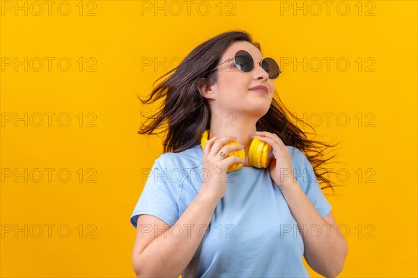 Studio portrait with yellow background of a caucasian woman moving her long hair while holding headphones on neck