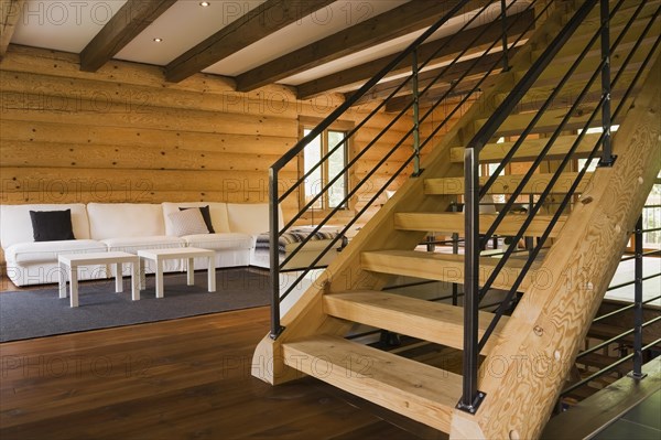 Wooden log stairs with black wrought iron railings and white cotton cloth upholstered sofas in living room inside contemporary style log home, Quebec, Canada, North America