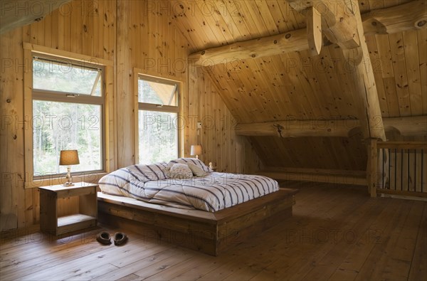 King size bed with wooden frame in master bedroom on upstairs floor inside handcrafted Eastern white pine Scandinavian log cabin home, Quebec, Canada, North America