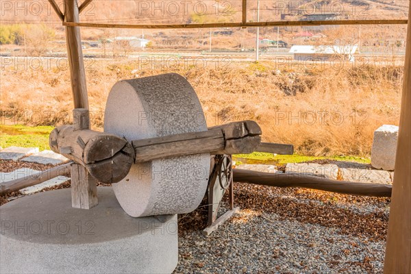 Old antique millstone and wheel in covered shelter with mountain community in background in South Korea