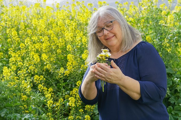 Smiling mature woman in her sixties with white hair and glasses holding a bouquet of daisies in her hands with a field with flowers in the background