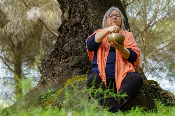 Mature woman with white hair meditating in the forest with her eyes closed and a Tibetan singing bowl ringing