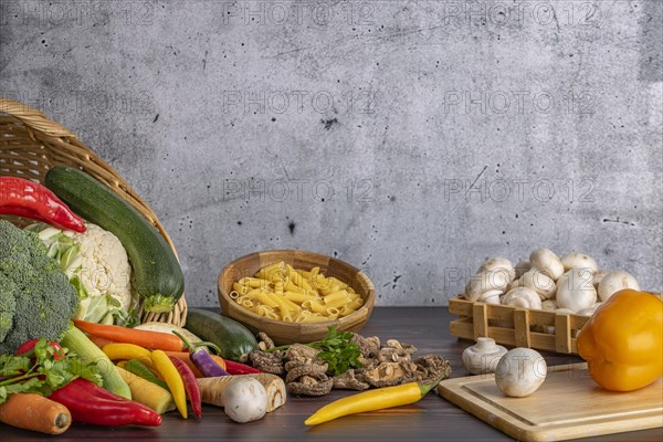 Various fresh vegetables such as peppers, cauliflower, peppers, mushrooms and broccoli around and in a basket with a bowl of pasta next to it on a wooden surface