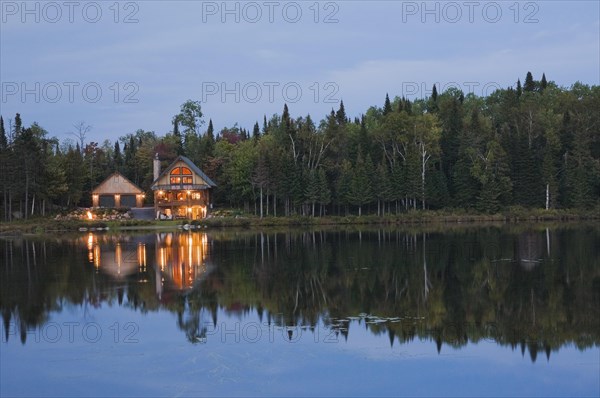 Illuminated two car garage and handcrafted two story spruce log cabin home with fieldstone chimney and green sheet metal roof on edge of lake at dusk in late summer, Quebec, Canada, North America