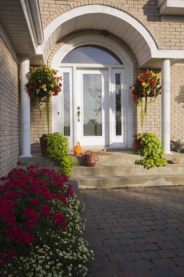 Front entrance door on tan brick with white trim portico and raised border with red Pelargonium, Geranium flowers in summer, Quebec, Canada, North America