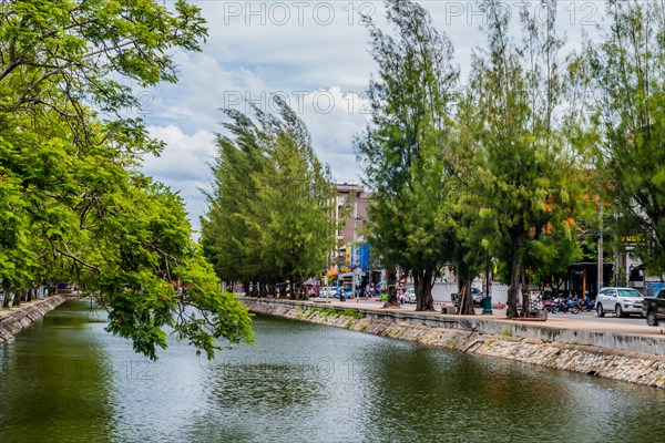 Cloudy sky reflecting on canal water, with a vibrant urban tree-lined waterfront, in Chiang Mai, Thailand, Asia