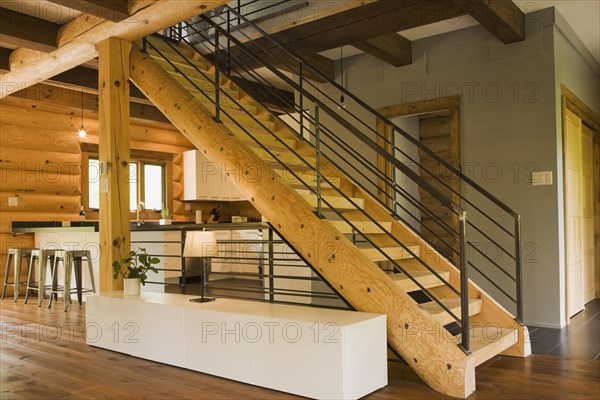 Kitchen with white wooden cabinets, metal bar stools and wooden log stairs with black wrought iron railings and long cabinet table inside contemporary style log home, Quebec, Canada, North America