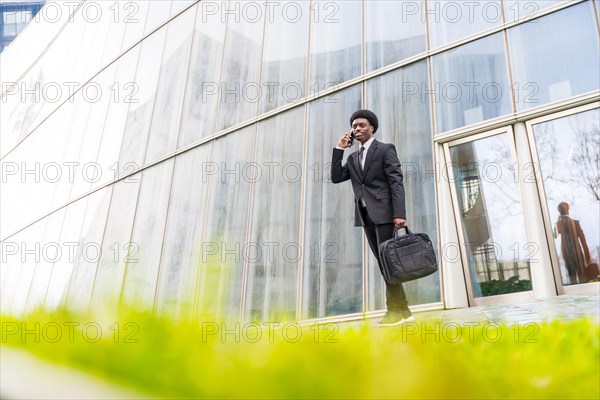 Wide view full length photo of a young african busy man coming out the office talking to the mobile