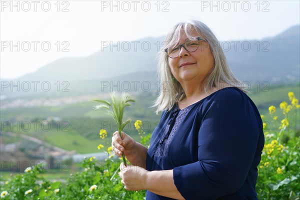 Older white-haired woman in the field with ears of barley in her hands with mountain scenery in the background