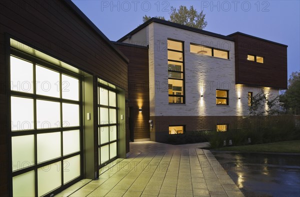 Illuminated two car garage and beige stone with brown cedar wood siding modern cubist style home facade with paving stone and black asphalt driveway at dusk in summer, Quebec, Canada, North America