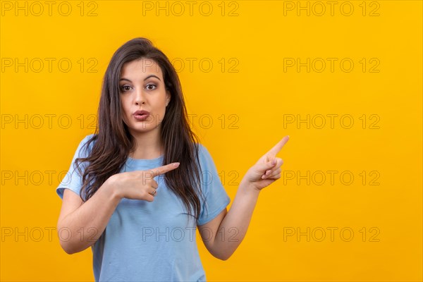 Studio portrait with yellow background of a surprised woman looking at camera and pointing aside to a blank space