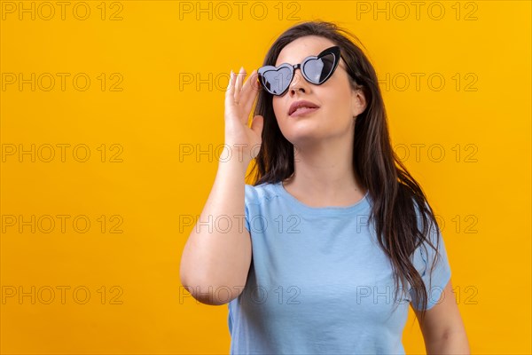 Studio portrait with yellow background of a chic woman with heart shape sunglasses looking up