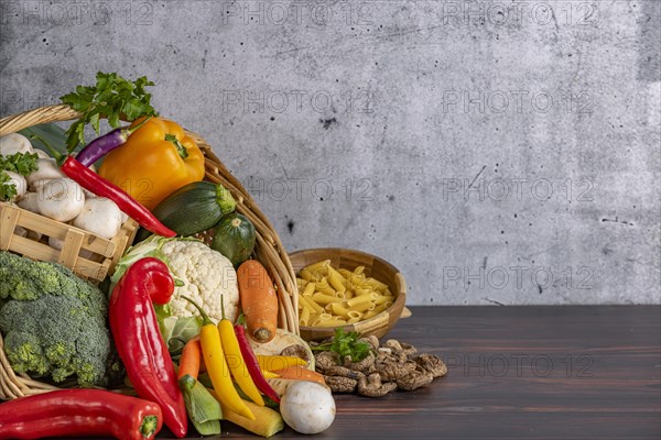 Various fresh vegetables such as peppers, cauliflower, peppers, mushrooms and broccoli in a basket with a bowl of pasta next to it on a wooden surface