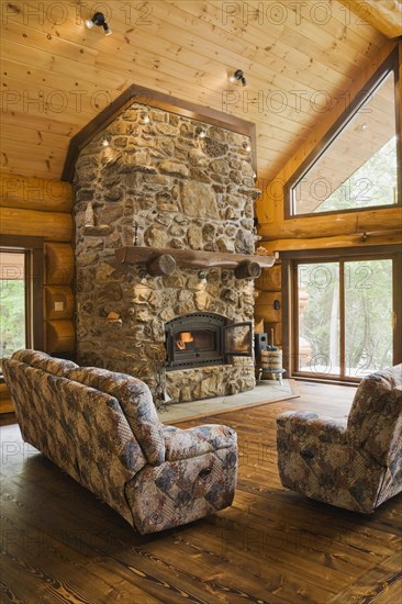 Upholstered sofas with lit fieldstone and porous rock fireplace in living room inside handcrafted red cedar log cabin home, Quebec, Canada, North America