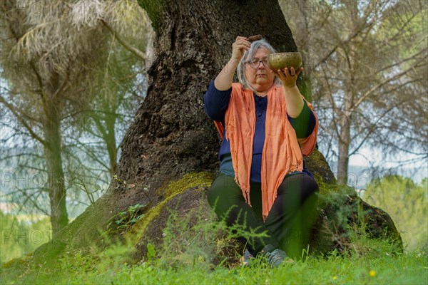 Mature woman with white hair meditating in the forest with her eyes closed and a Tibetan singing bowl ringing